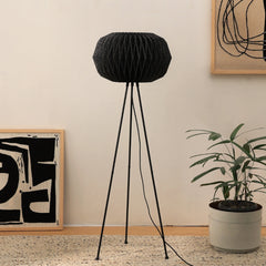 A modern origami-inspired floor lamp with a black linen shade on a tripod stand, standing in a room with decorative plants and wall art.