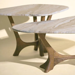 Two maya tables with oval light beige marble tops and sculptural curved wooden bases, shown on a warm cream background.