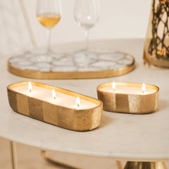 Fleur Votive- Two lit candles in gold candle holders on a table with glasses in the background