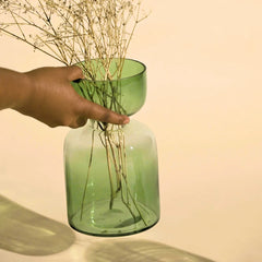 Hand holding a sage green tinted glass vase with dried plants against a beige background