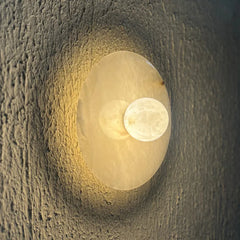 Modern alabaster wall-mounted light fixture featuring a large oval frosted glass shade and a smaller circular moon-shaped glass bulb, casting a warm yellow glow against a textured dark gray wall.