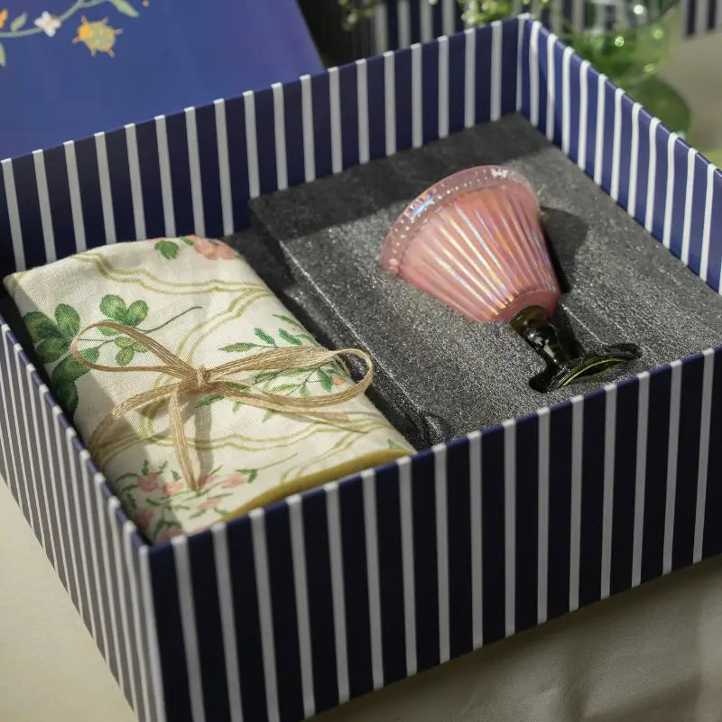 A Cushion Cover and a pink Votive Candle with a flared rim, nestled in gray foam packaging inside a striped box.