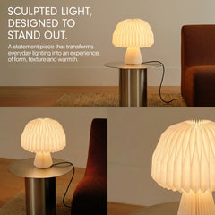 A sculptural pleated Mushroom Origami table lamp shown in a styled interior setting. The lamp has a warm white ribbed shade and base and is lit, casting a soft glow.
