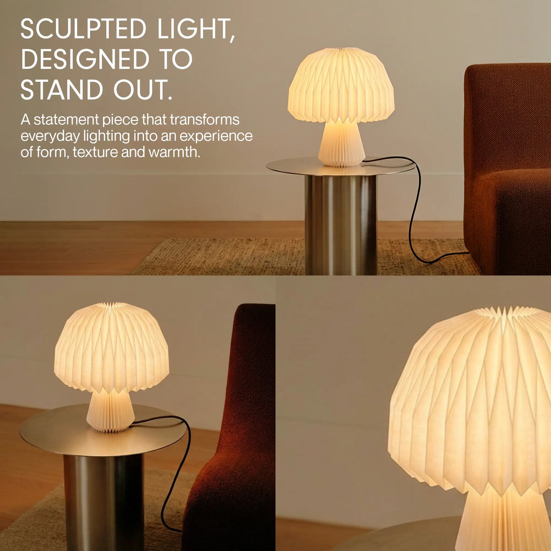A mushroom-shaped table lamp with a pleated white paper-like shade and base, glowing warm yellow on a wooden cabinet in a styled interior setting.