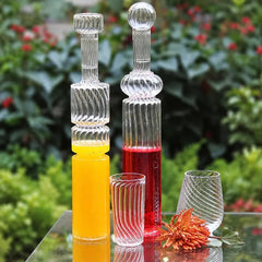 Firdaus two clear ribbed glass decanters with lid and matching small tumblers, shown on a glass tabletop with liquid samples in one bottle each.