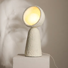 A handcrafted Echo Sphere Table Lamp made from paper mache, with a textured, beige surface and a round top, casting a warm glow.