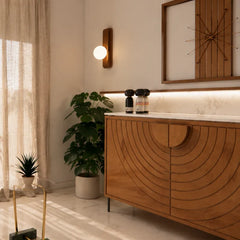 A modern wooden bathroom vanity cabinet with a curved slatted front and marble countertop, styled in a warm, sunlit bathroom with a potted plant, wall sconce, and framed wall art.