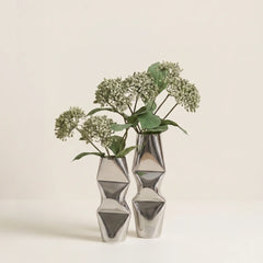 Two metallic silver Chrome Dented Vases -like candle holders with a faceted, twisted geometric design, styled with dried green and white flowers.