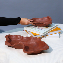 Two Kai organic bowls in brown ceramic leaf-shaped design, one on the table and other person holding over a stack of books.
