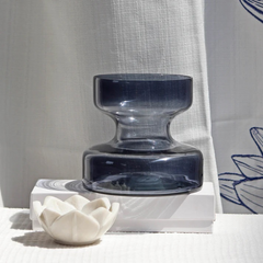Decorative slate grey glass vase on a white surface with a white ceramic flower and a white curtain in the background