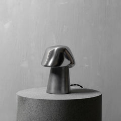 A mushroom-shaped table lamp with a glossy metallic silver cap and a matte dark gray base, displayed on a cylindrical gray pedestal against a textured light gray wall.
