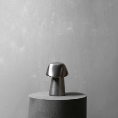 A small metallic mushroom-shaped object with a brushed silver cap and darker stem, displayed on a cylindrical pedestal against a textured light gray wall.