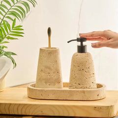 Eara Soap Dispenser Set - a beige travertine stone bathroom set on a wooden tray, featuring a toothbrush holder & a matching pump bottle with a black dispenser, with a hand reaching for the pump.