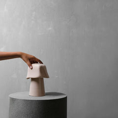A hand holds a small sculptural ceramic object with a wide rounded top and narrow cylindrical base, placed on a round gray pedestal against a textured light gray wall.