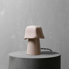 A minimalist mushroom-shaped table lamp in a matte blush pink ceramic finish, displayed on a textured dark gray cylindrical pedestal against a light gray plaster wall.