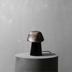 A mushroom-shaped table lamp with a metallic bronze cap and a black cylindrical base, displayed on a round gray pedestal against a textured gray wall.