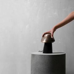 A mushroom-shaped table lamp with a metallic bronze dome and a black base, shown on a textured gray pedestal with a person's hand reaching toward the top.