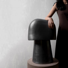 A modern mushroom-shaped black stool with a smooth rounded cap and tapered pedestal base, shown on a round pedestal against a light gray wall, with a woman in a dark dress partially visible on the right.