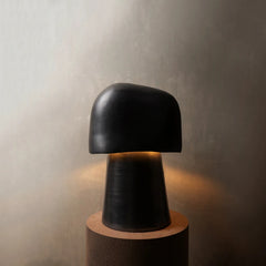 A modern black ceramic table lamp with a rounded dome-shaped shade and a tapered base, illuminated from within to cast a warm glow, set on a cylindrical wooden pedestal against a textured gray wall.