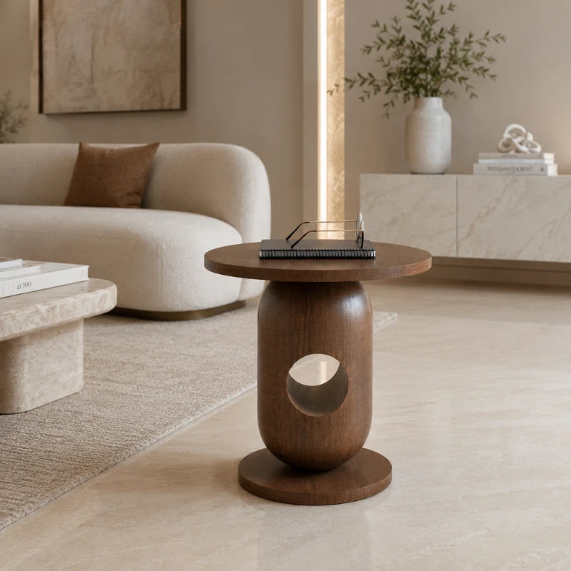 A round wooden end table with a sculptural cylindrical base and a circular cutout, styled in a modern living room setting.