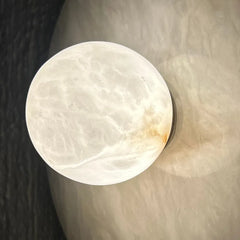 Halo wall sconce -round, polished white alabaster stone or crystal sphere with natural marbled veining and a small warm brownish patch near the lower right edge, resting on a textured beige surface.