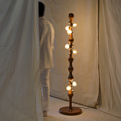 A tall, twisted wooden floor lamp with multiple warm glowing globe bulbs, standing on a circular base against a draped cream fabric backdrop.