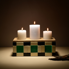 Three white candles on a decorative Blade of Grass Candle Holder made of wooden block with a checkered pattern against a dark background. 