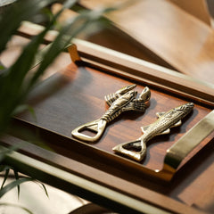Two gold brass fish-shaped bottle openers on a wooden surface with a plant in the foreground.