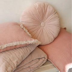 A soft blush pink bedding set styled with a round tufted pillow, a folded quilted blanket, and a matching pillow with lace trim, set against a neutral cream background.