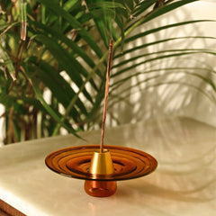 Amber-colored incense holder with a single stick on a marble surface with greenery in the background