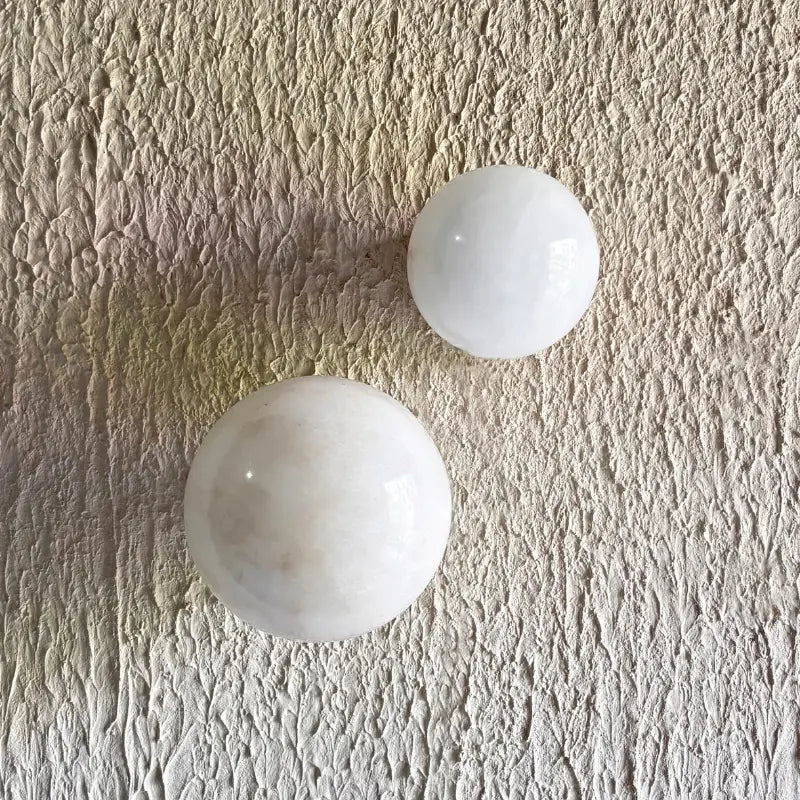 Two smooth white polished alabaster stone sphere wall sconces resting on a textured beige surface.