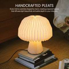 A mushroom-shaped table lamp made of pleated paper, glowing warm white on a stack of books. Text at the top reads 'HANDCRAFTED PLEATS' with descriptive copy about layered texture and interior lighting.