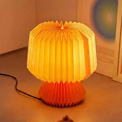 Decorative pleated table lamp with a mushroom-shaped design, glowing with a warm yellow light. The lamp features a pleated white upper dome and a pleated orange lower base, sitting on a wooden surface.