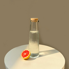 Clear glass bottle with cork lid on a round white surface with a half-cut grapefruit, against a beige background.