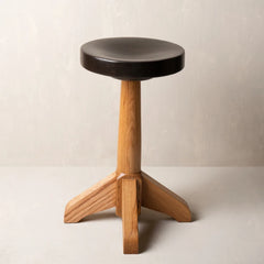 A rustic wooden stool with a round black seat cushion and a natural oak three-legged base, set against a neutral light.