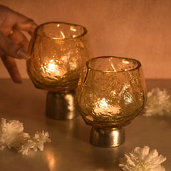 Two lit glass candle holders with Sandcast metal base on a reflective surface with flowers around, warm lighting.