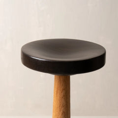 A modern stool with a round black seat and a natural wood leg, shown against a neutral light gray textured background.