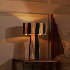 Wald Table Lamp with striped black and natural raffia design on a wooden table in a warm-lit room.