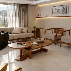 A modern living room featuring a mid-century modern furniture set, including a beige fabric sofa, two wooden armchairs with beige cushions, a low oval wooden coffee table, and a round wooden side table. The room has warm beige walls, a built-in floating shelf with warm lighting, and a large window with sheer curtains.