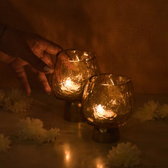 Two lit glass tealight candle holders with Sandcast metal base on a wooden surface with a warm glow, surrounded by white flowers.