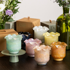 A set of six ribbed glass candle holders with scalloped rims in assorted pastel and warm tones, displayed on a dark wooden surface with decorative flowers, Doft gift boxes.