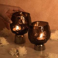 Two lit candle holders with sandcast metal base and a hand holding one, surrounded by white flowers on a soft surface.