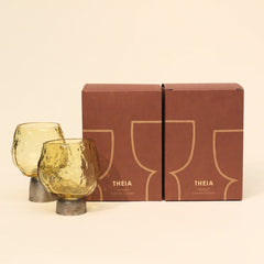 Two yellow glass candle holders with Sandcast metal base placed along a matching brown box featuring 'THEIA' branding on a beige background.