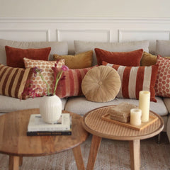 A styled living room scene with a beige sofa covered in multiple decorative throw pillows in rust and Cuddle Sand Velvet Round Cushion, striped, and patterned designs. 
