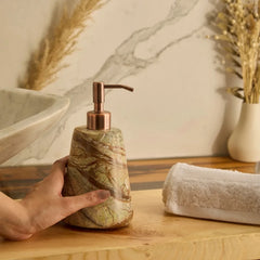 A hand holds a marble-look Reen soap dispenser with a copper pump on a wooden bathroom counter next to a white towel, with a marble sink and dried grass in the background.