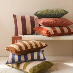 A stack of Holly Eucalyptus Velvet Stripe Cushion Covers in various colors including brown, blue, green, orange, and burgundy, arranged on a wooden bench and the floor against a neutral beige wall.