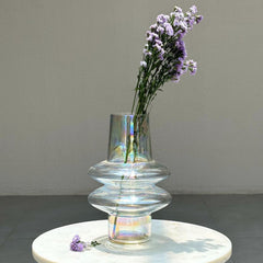 Glass vase with iridescent finish holding purple flowers on a white surface.