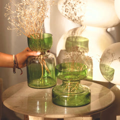 Sage Green glass vase with dried branches on a table, with a hand holding another similar vase. Two different sizes.