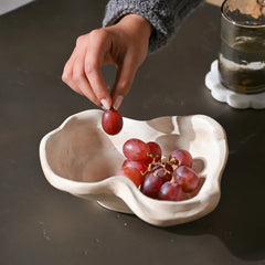 Kai Organic Bowl Blush Sandstone, organic-shaped off-white ceramic bowl filled with red grapes, set on a dark tabletop next to a glass of water.