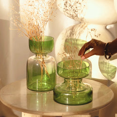 Two sage green tinted glass vases with dried plants on a round table.